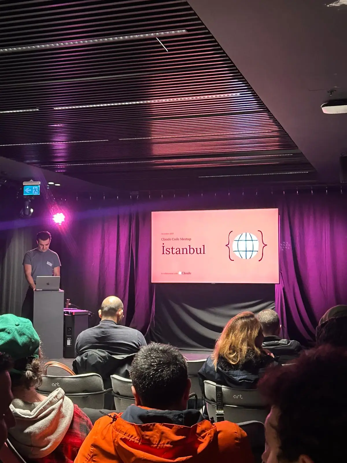 Prezentare pe scena principală a Istanbul Claude Summit 2025 cu Claude Code Meetup Istanbul afișată pe ecran, iluminare ambientală violet, vorbitor la podium cu laptop și public implicat care urmărește sesiunea de deschidere la evenimentul pentru dezvoltatori Anthropic din Turcia